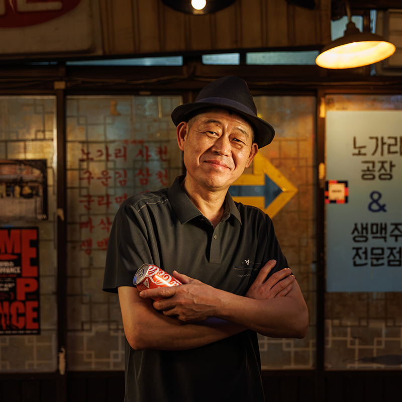 신흥시장 노가리공장 정헌주 사장이 가게 앞에서 코카-콜라 캔을 들고 팔짱을 낀 채 카메라를 바라보는 모습.