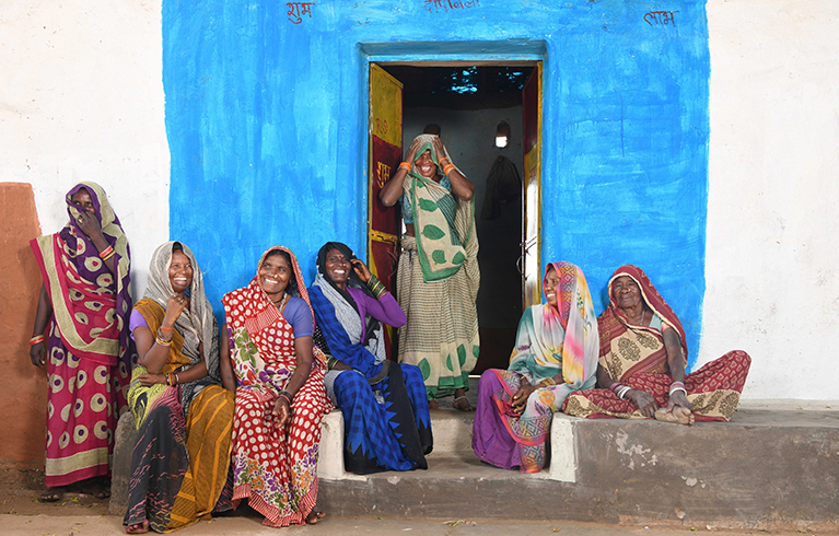 7 women in a row, some sat some standing, against a wall and blue doorway