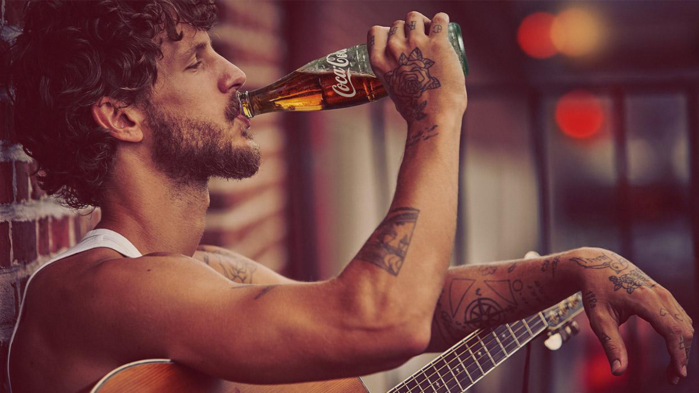 Un bărbat tânăr, cu barbă, ține o chitară și bea Coca-Cola dintr-o sticlă