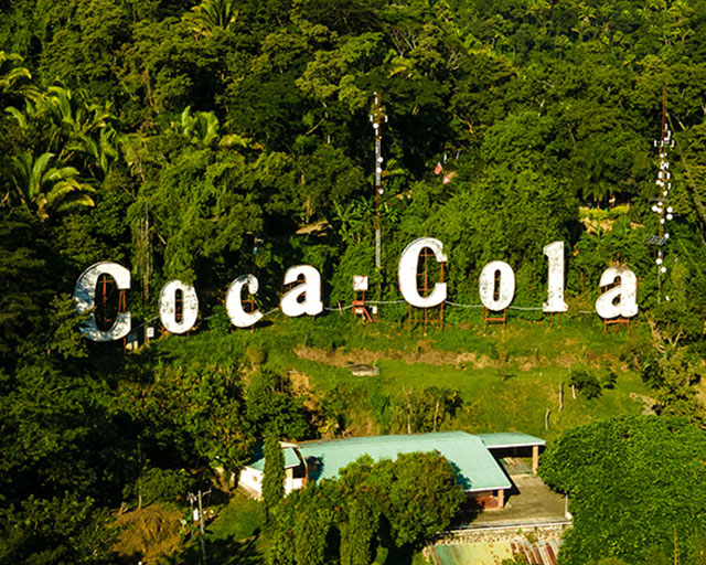 Casă într-un luminiș în junglă pădure, la marginea luminișului sunt panouri cu litere care formează cuvintele Coca Cola.