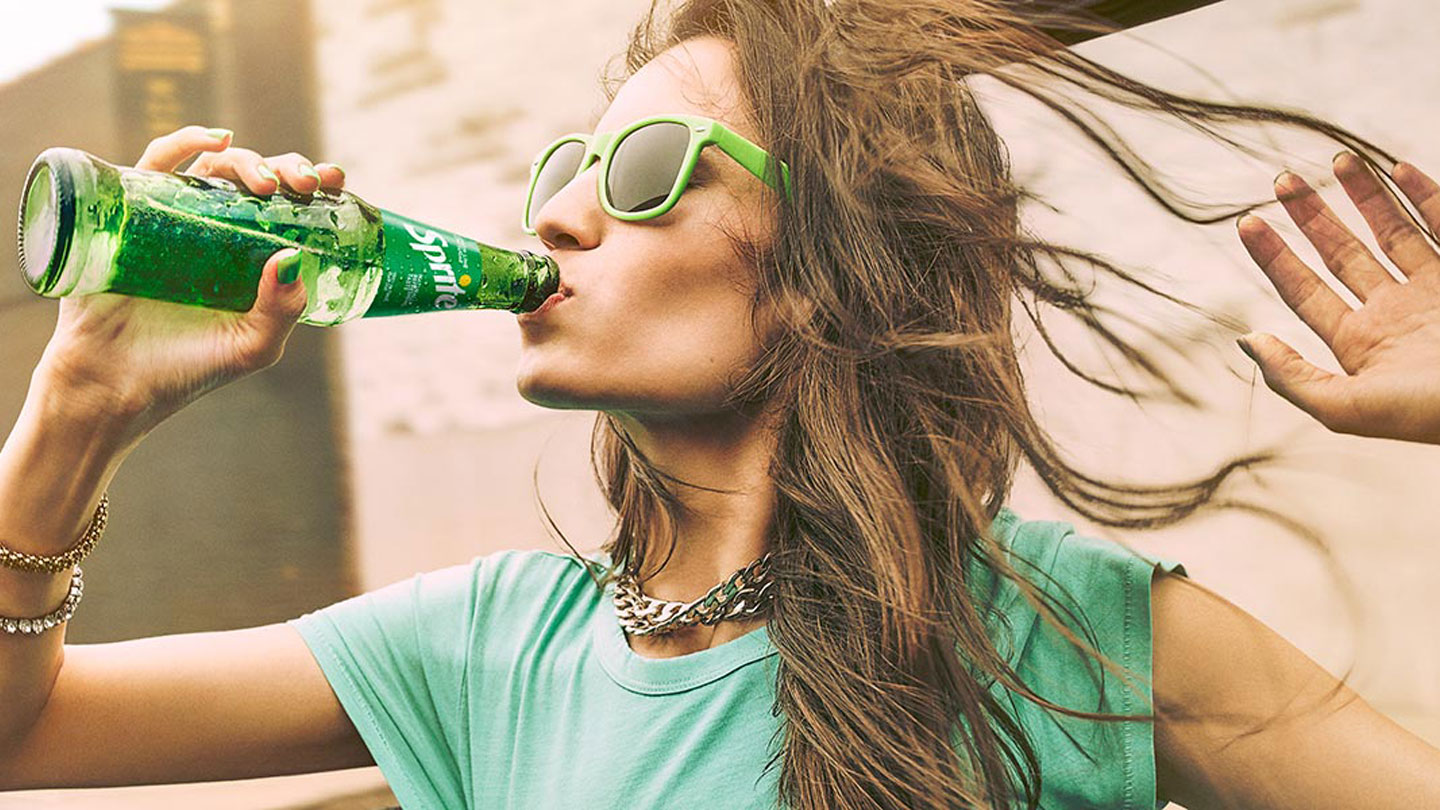 Đevojka u zelenoj majici i zelenim naocarima pije Sprite iz flašice