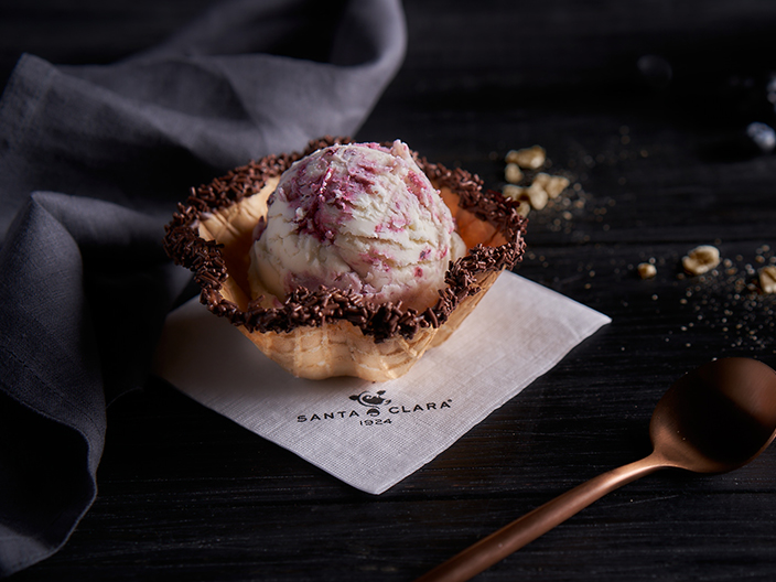 Una bola de helado de vainilla con vetas de frambuesa o fresa, servida en un tazón de waffle con borde de chocolate y chispas. El tazón descansa sobre una servilleta blanca con el logo "SANTA CLARA" en la esquina inferior izquierda. A la derecha, se ve una cuchara de cobre y algunas nueces esparcidas sobre una superficie oscura.
