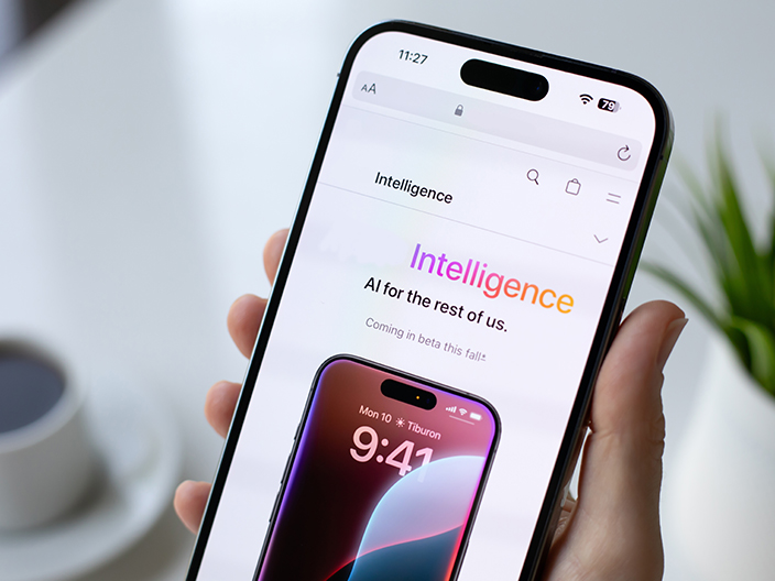 Una mano sostiene un teléfono inteligente moderno con una pantalla que muestra la palabra "Intelligence" en colores vibrantes y una imagen de otro teléfono con la hora "9:41". En el fondo, se ve una taza de café y una planta verde.