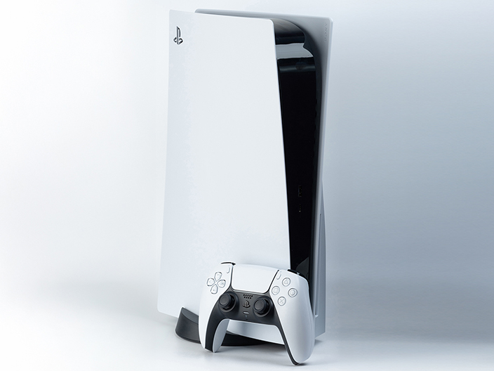 Una consola de videojuegos PlayStation 5 blanca y negra, con su diseño vertical y curvo, se encuentra de pie sobre una superficie clara. Un control DualSense blanco a juego está apoyado en la base de la consola. El fondo es de un color gris claro y uniforme.