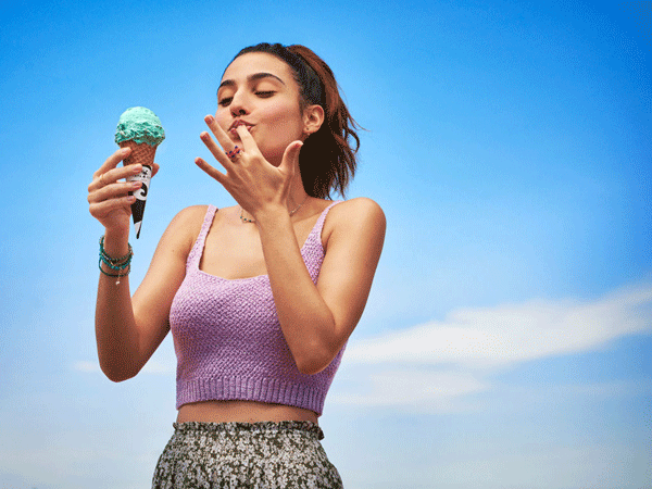 Una mujer disfruta de un helado de menta bajo un cielo azul brillante. Lleva una blusa lila y una falda floreada. Lambe el helado de sus dedos.