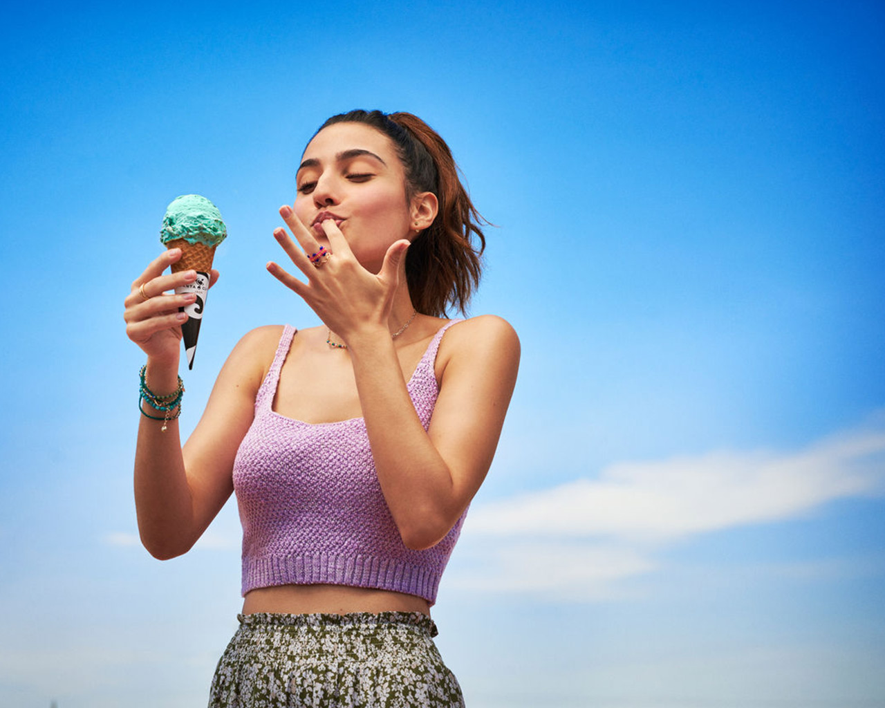 Una mujer disfruta de un helado de menta bajo un cielo azul brillante. Lleva una blusa lila y una falda floreada. Lambe el helado de sus dedos.