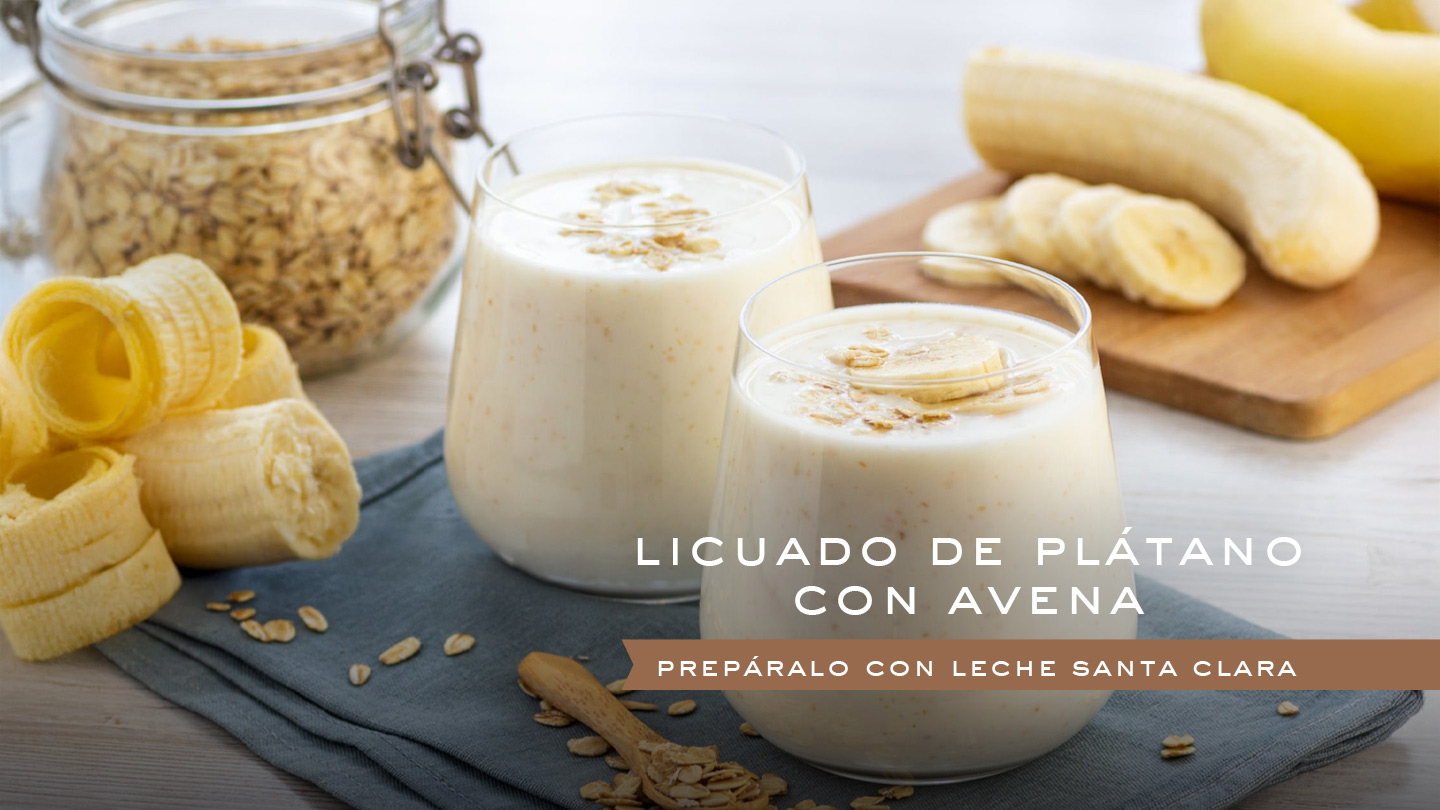 Dos vasos de licuado de plátano con avena, decorados con rodajas de plátano y avena espolvoreada, sobre una tela azul. Al fondo, se ve un frasco de avena, un plátano entero y rodajas de plátano en una tabla de madera. En la parte superior de la imagen, un texto dice "LICUADO DE PLÁTANO CON AVENA" y debajo "PREPÁRALO CON LECHE SANTA CLARA".