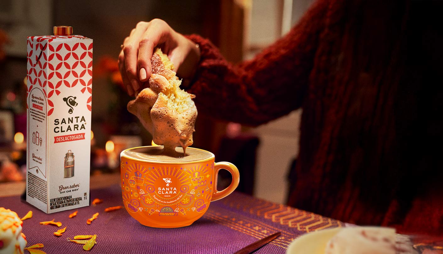 Una persona sumerge un pan dulce en una taza naranja de chocolate caliente, junto a un cartón de leche Santa Clara.