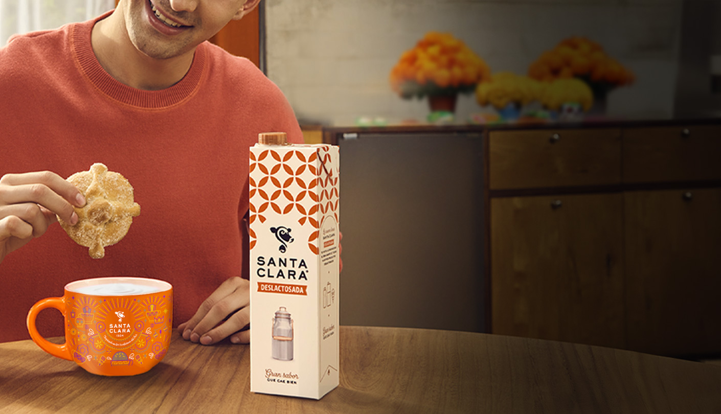 Un primer plano de una persona sumergiendo una galleta en una taza naranja, con un cartón de leche Santa Clara al lado.