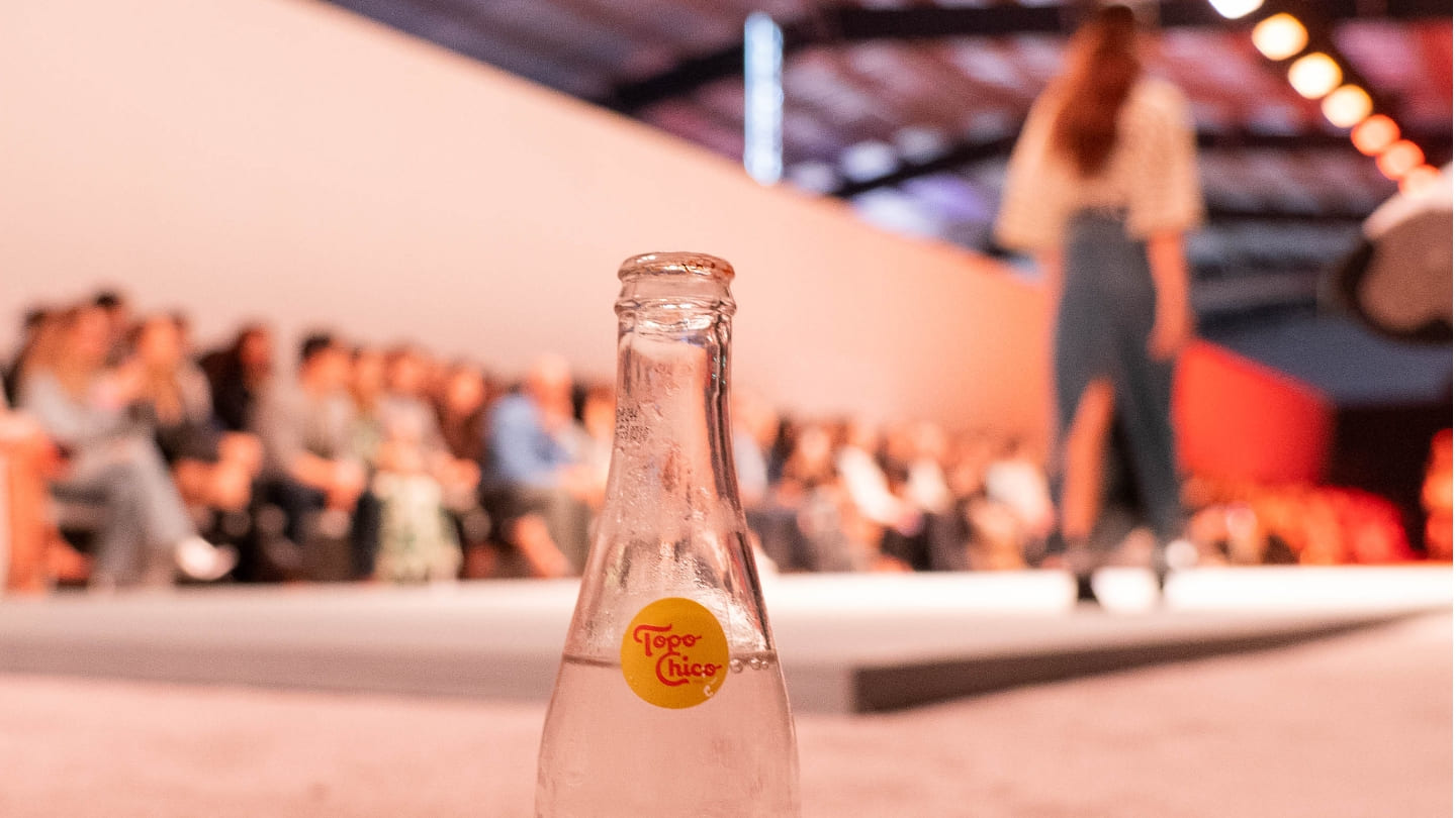 Botella de Topo Chico. De fondo se observa personas sentadas mirando hacia una pasarela de moda.