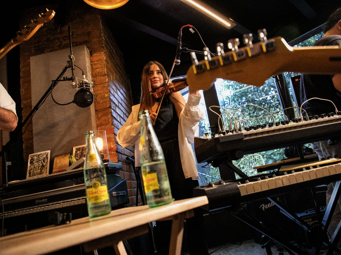 Una joven toca el violín. Delante de ella, se observan dos botellas de Topo Chico sobre una mesa.