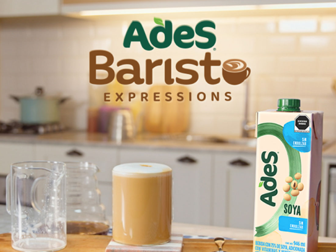 Vaso de café con espuma sobre una servilleta junto a jarros de café y un envase de leche de soya AdeS sobre la mesa de una cocina y por encima se encuentra el logo de AdeS Barsita Expressions 