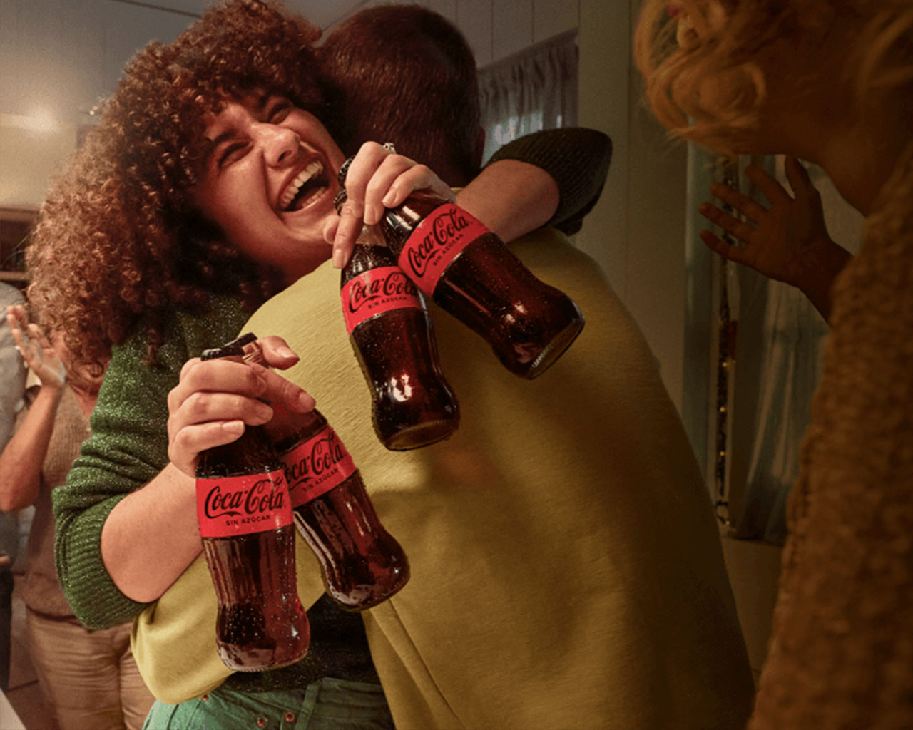 Mujer de pelo ondulado riendo abrazando a un hombre mientras sostiene dos botellas de coca en cada mano.