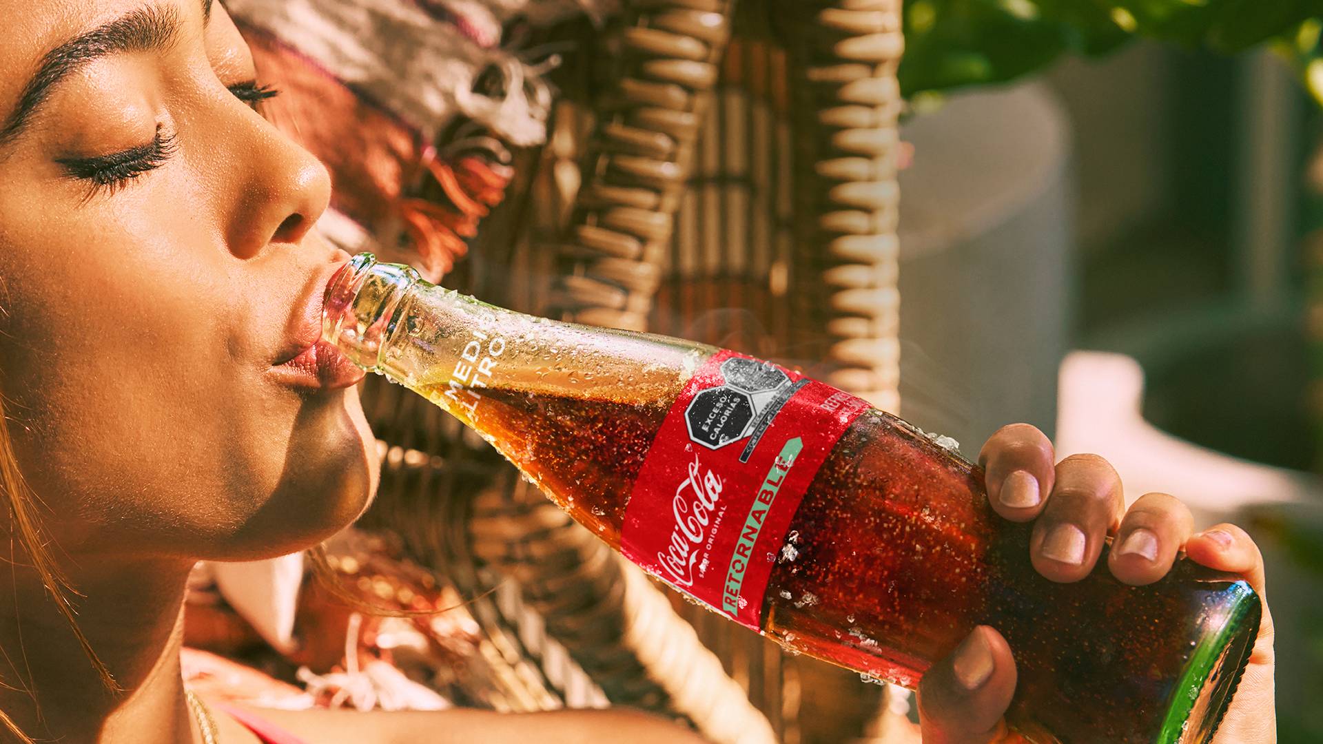 Una mujer bebiendo de una botella de Coca-Cola de vidrio helado en un día soleado, mientras el fondo muestra un tejido de mimbre y algo de vegetación.