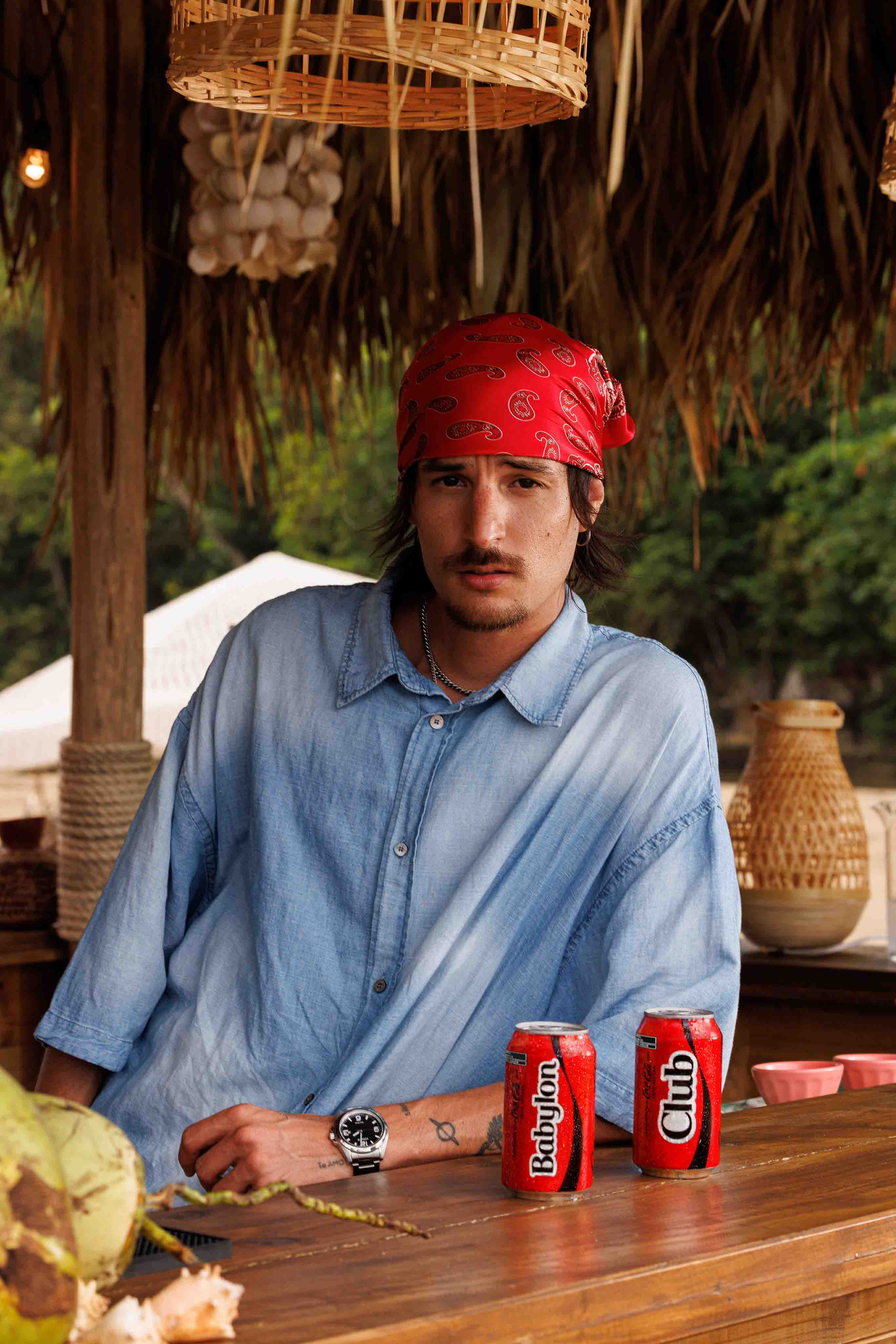Danny Ocean con bandana roja y camisa sentado en un bar de playa con dos latas de Coca-Cola. Una tiene inscripta la palabra "Babylon", y la otra "Club".