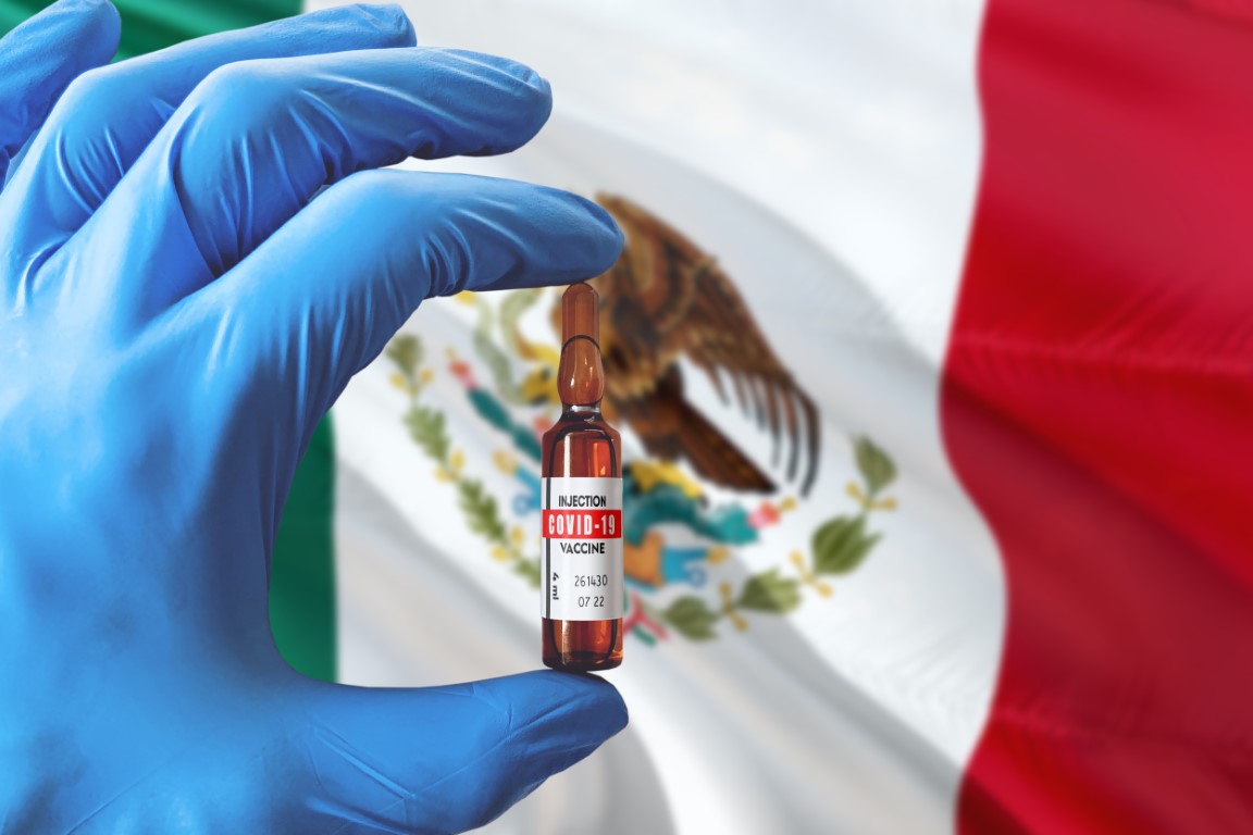 Manos con guantes sujetando una vacuna con bandera de México de fondo