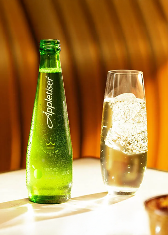 Bouteille verte Appletiser à côté d’un verre rempli, posée sur une table dans une ambiance chaleureuse et lumineuse.