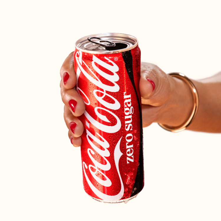 Une main avec des ongles vernis rouges et un bracelet en or tient une canette Coca‑Cola Zero Sugar bien fraîche, avec des gouttelettes de condensation visibles.