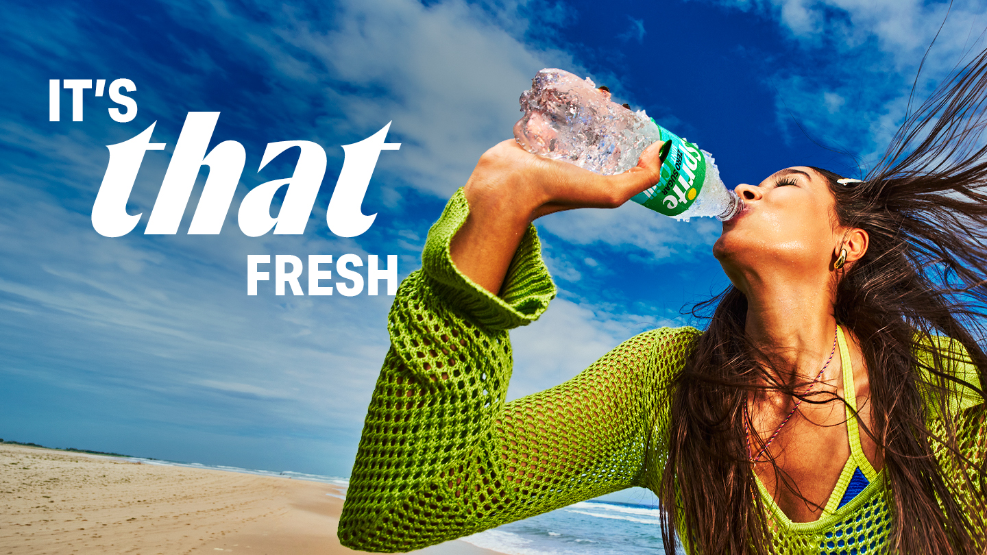 Advertentiebeeld van een vrouw op het strand die uit een fles Sprite drinkt. Ze draagt een felgroene gehaakte trui en haar haar waait in de wind. Op de achtergrond zijn een blauwe lucht met wolken en de zee zichtbaar. Links in beeld staat de witte tekst: “IT’S that FRESH”.
