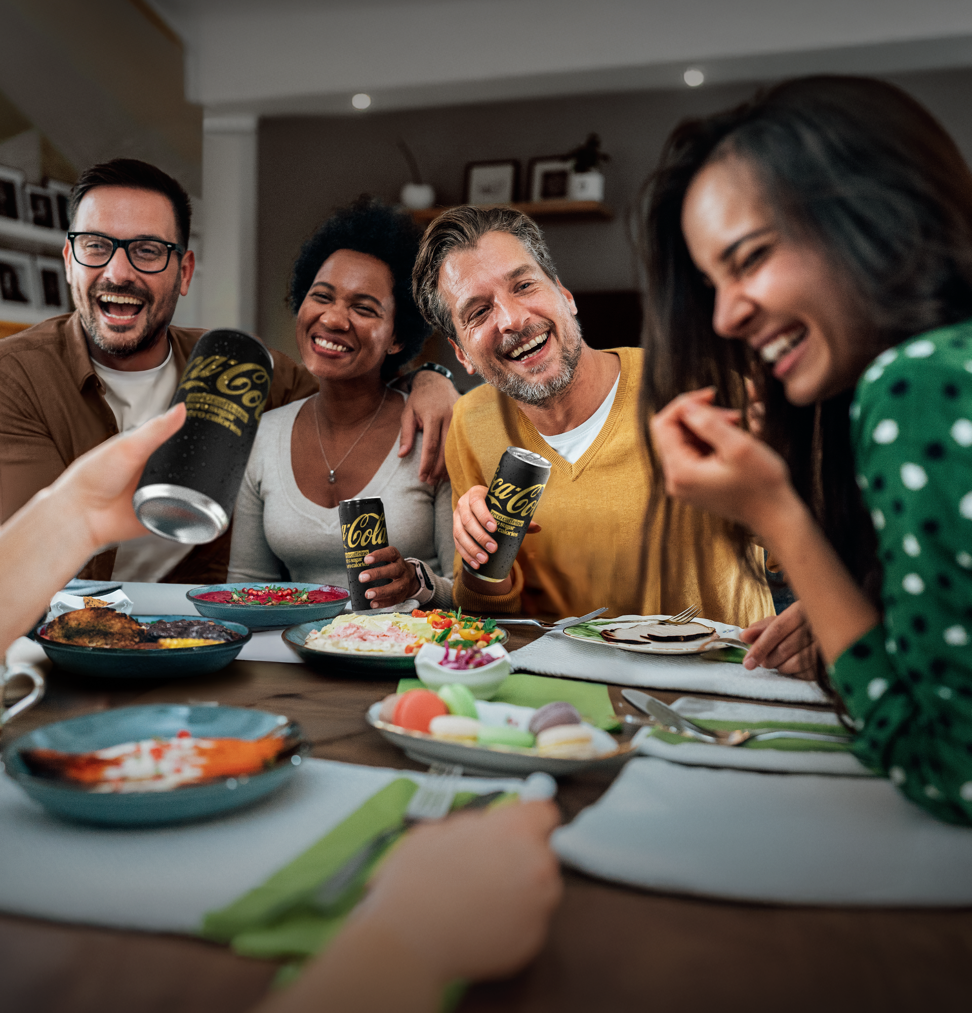 Groep vrienden aan tafel met eten en blikjes Coca‑Cola Zero, in een gezellige setting.