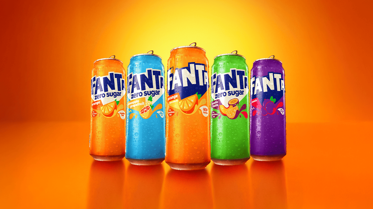 Vijf Fanta-blikjes staan naast elkaar op een oranje achtergrond. De blikjes hebben verschillende kleuren en smaken: oranje, blauw (zero sugar), oranje, groen (zero sugar) en paars. Op elk blikje staan fruitillustraties en er zitten waterdruppels op, wat een frisse uitstraling geeft.