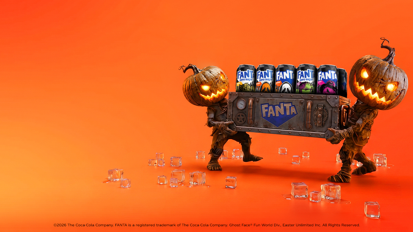 Twee figuren met verlichte pompoenhoofden dragen een metalen krat met verschillende blikjes Fanta. De scène heeft een oranje achtergrond met verspreide ijsblokjes en een Halloween-thema.