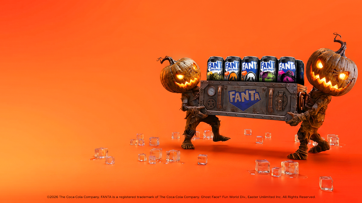 Twee figuren met verlichte pompoenhoofden dragen een metalen krat met verschillende blikjes Fanta. De scène heeft een oranje achtergrond met verspreide ijsblokjes en een Halloween-thema.