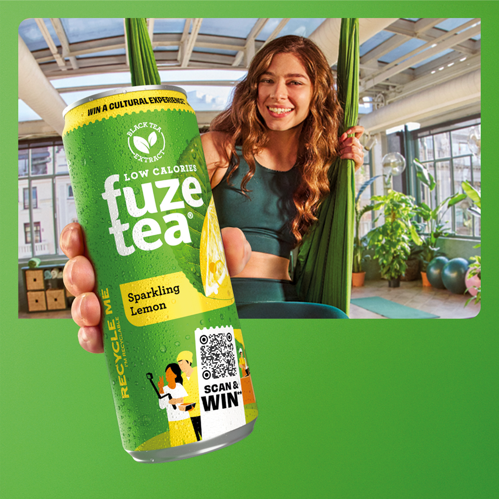 Een persoon staat in een lichte yogastudio en houdt een fles Fuze Tea dicht bij de camera. De persoon draagt groene sportkleding en staat op een yogamat. Op de achtergrond zijn kamerplanten, yogamatten en sportaccessoires te zien; het gezicht is onherkenbaar gemaakt.