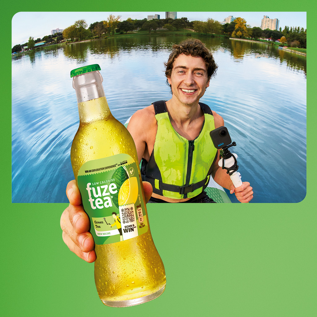 Een persoon zit in een kajak op rustig water en houdt een fles Fuze Tea dicht bij de camera. De persoon draagt een felgroen reddingsvest en sportkleding en peddelt met een kajakpeddel. Het gezicht is onherkenbaar gemaakt en op de achtergrond is water en natuur te zien.