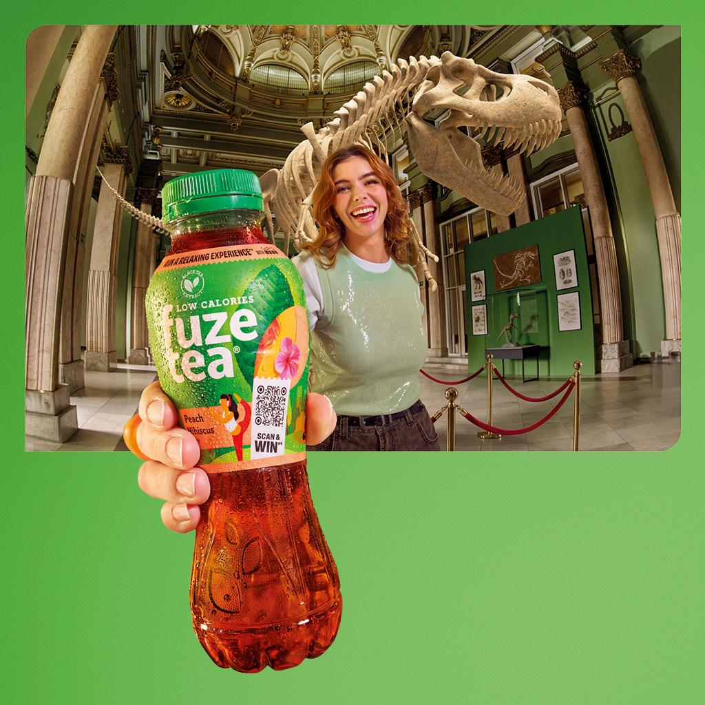 Een persoon staat in een museumzaal en houdt een fles Fuze Tea dichtbij de camera. Op de achtergrond is een groot dinosaurus‑skelet te zien, met vitrines en afzetlinten in de ruimte. De persoon draagt een groen shirt en spijkerbroek; het gezicht is onherkenbaar gemaakt.