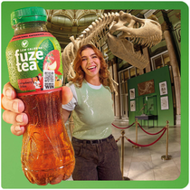 Een persoon staat in een museumzaal en houdt een fles Fuze Tea dichtbij de camera. Op de achtergrond is een groot dinosaurus‑skelet te zien, met vitrines en afzetlinten in de ruimte. De persoon draagt een groen shirt en spijkerbroek; het gezicht is onherkenbaar gemaakt.