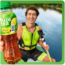 Een persoon zit in een kajak op rustig water en houdt een fles Fuze Tea dicht bij de camera. De persoon draagt een felgroen reddingsvest en sportkleding en peddelt met een kajakpeddel. Het gezicht is onherkenbaar gemaakt en op de achtergrond is water en natuur te zien.
