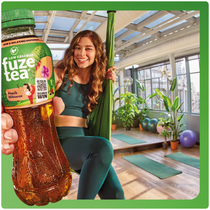 Een persoon staat in een lichte yogastudio en houdt een fles Fuze Tea dicht bij de camera. De persoon draagt groene sportkleding en staat op een yogamat. Op de achtergrond zijn kamerplanten, yogamatten en sportaccessoires te zien; het gezicht is onherkenbaar gemaakt.