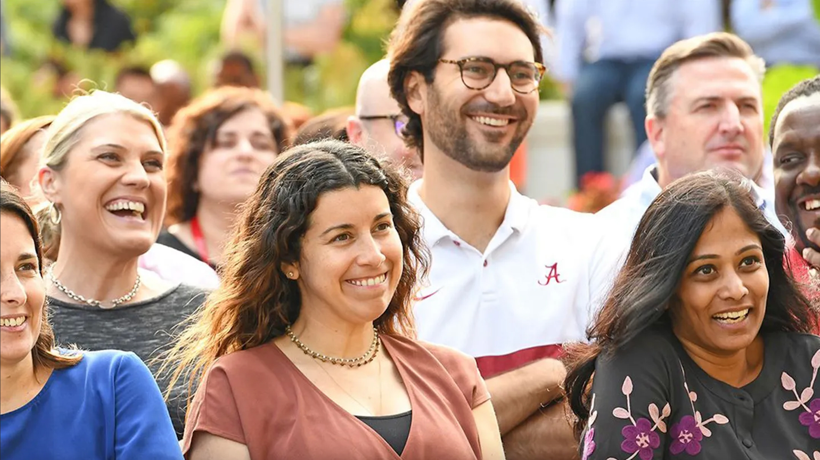 Een groep mensen zit dicht bij elkaar in een buitenomgeving met groen op de achtergrond. Ze dragen casual kleding, waaronder een blauwe blouse, een bordeauxrood T‑shirt, een witte poloshirt met een rood “A”-logo en een zwart shirt met bloemmotief. Enkele mensen lachen of kijken aandachtig naar iets buiten beeld. Het is zonnig en de sfeer oogt ontspannen en gezellig.