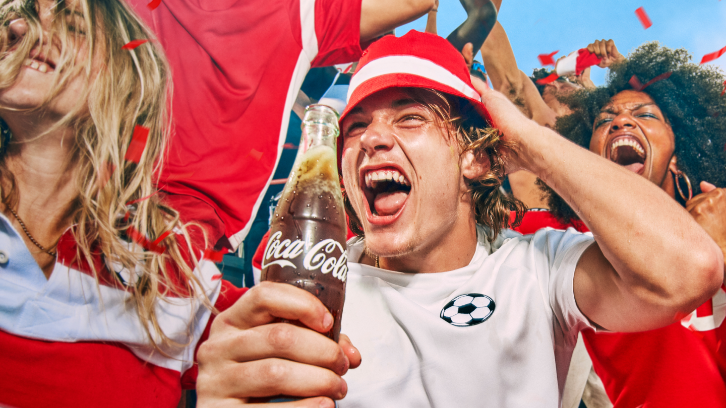 Hópur fólks í rauðum og hvítum fatnaði fagnar saman utandyra, með rauðu konfetti í loftinu. Einstaklingur heldur á Coca‑Cola flösku í forgrunni og annar drekkur úr flösku. Fótboltaþema, hátíðarstemning og stuðningur við íþróttaviðburð.