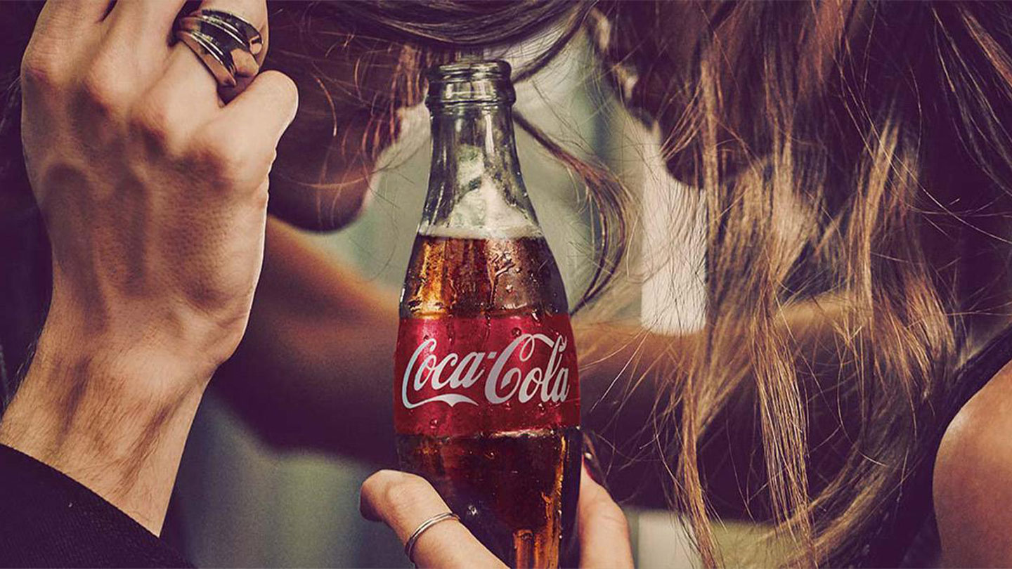 Bilda av et par som holder hodene sammen og som har en flaske Coca-Cola mellom seg. 