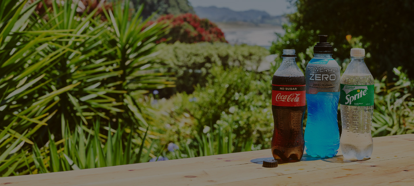 A bottle of Coca-Cola No Sugar, Powerade Zero, and Sprite No Sugar side by side in front of a garden