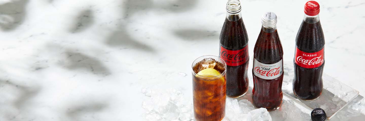 Coca-Cola bottles and a glass of Coca-Cola with ice on a surface exposed to the sunlight