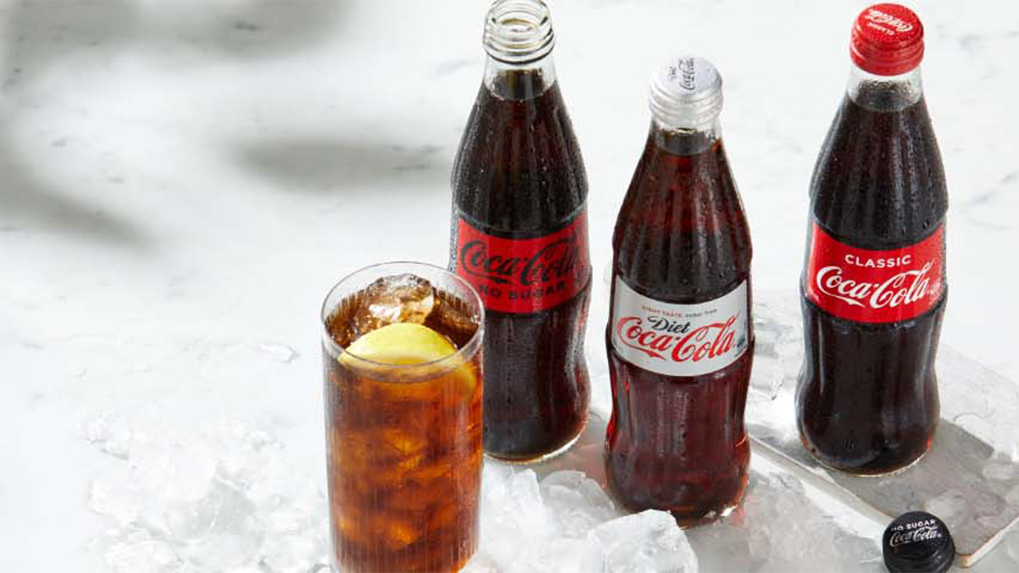 Coca-Cola bottles and a glass of Coca-Cola with ice on a surface exposed to the sunlight