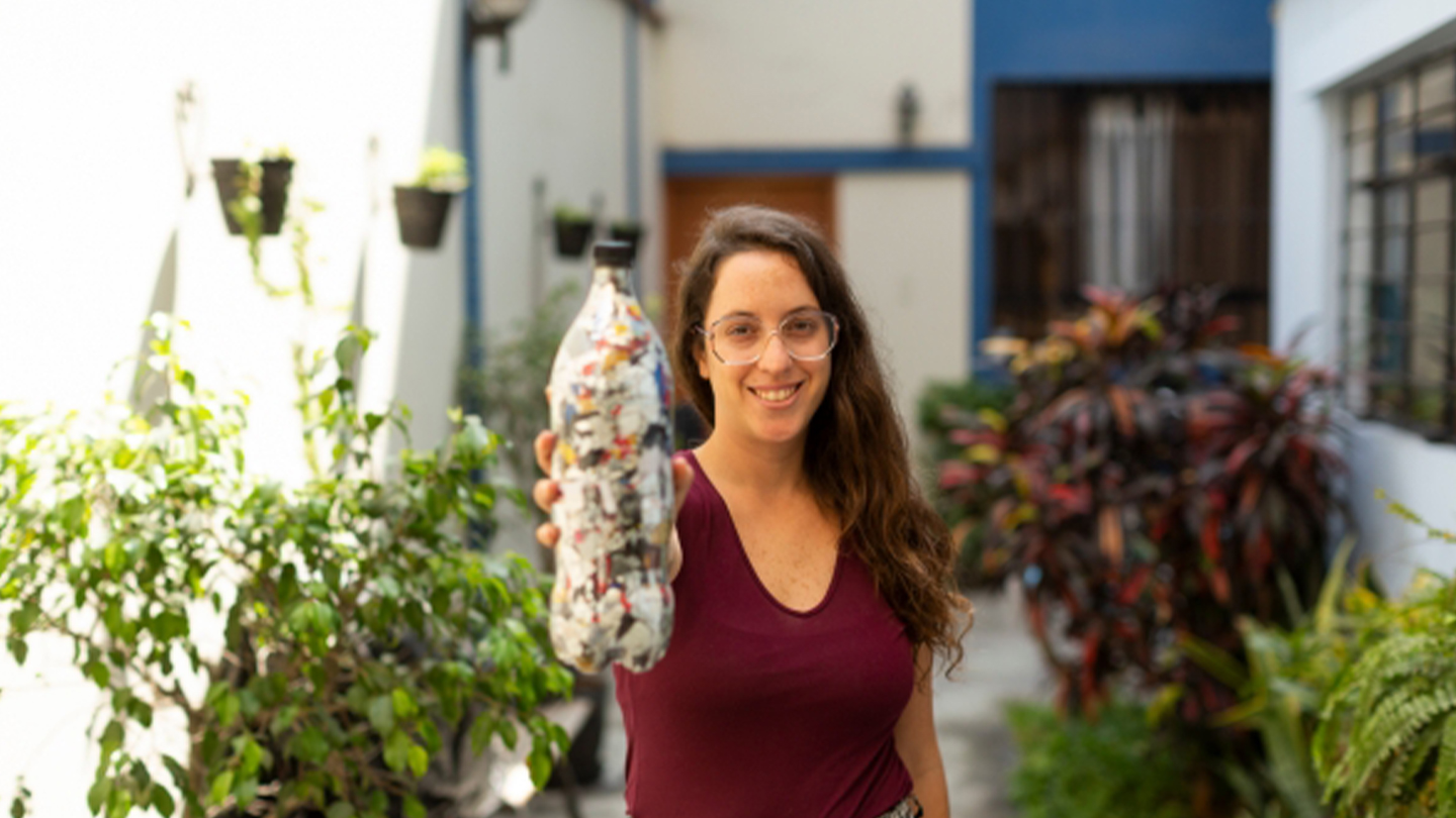 Chica sonriente sujeta una botella rellena de residuos plásticos en una terraza con plantas