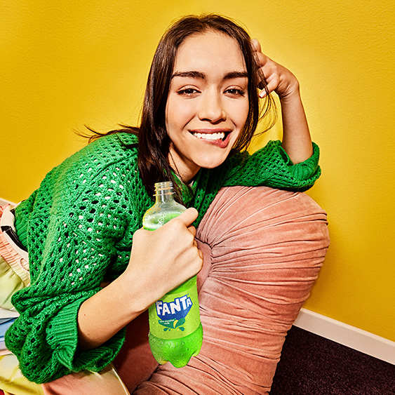 Una joven sonríe con una botella de Fanta Manzana, vestida con un suéter verde y recostada sobre un almohadón en una habitación amarilla.