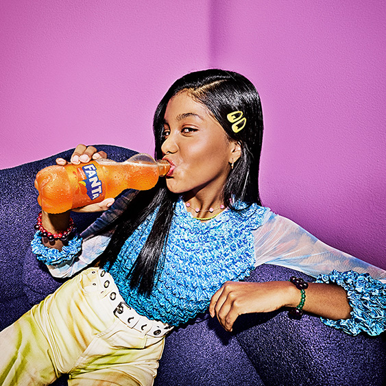 Mujer bebiendo de una botella de Fanta Naranja recostada en un sillón.