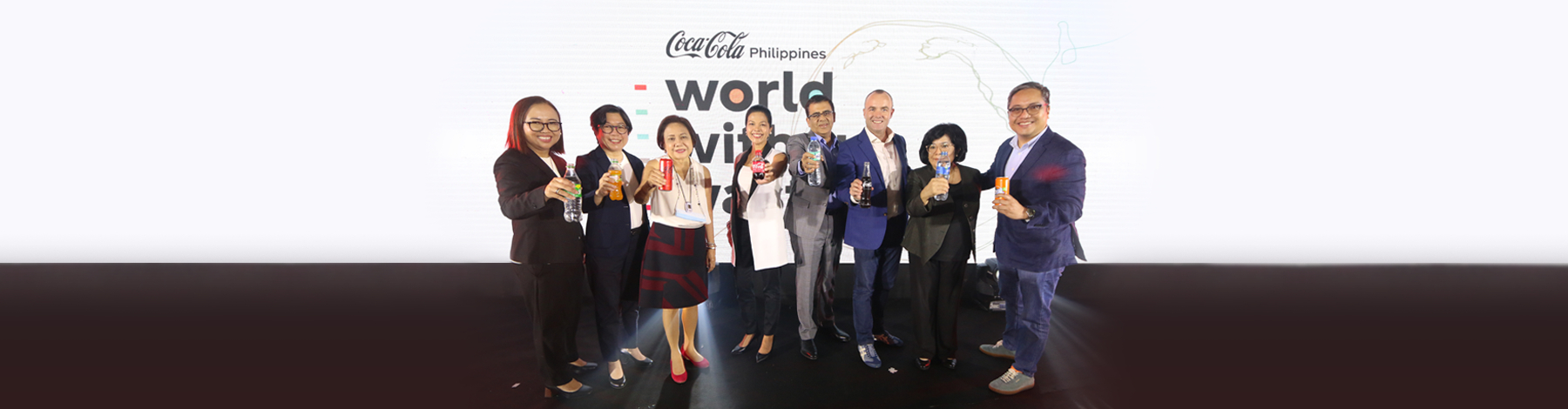 Group of people in suits holding Coca-Cola products in front of a Coca-Cola Philippines World Without Waste advertising
