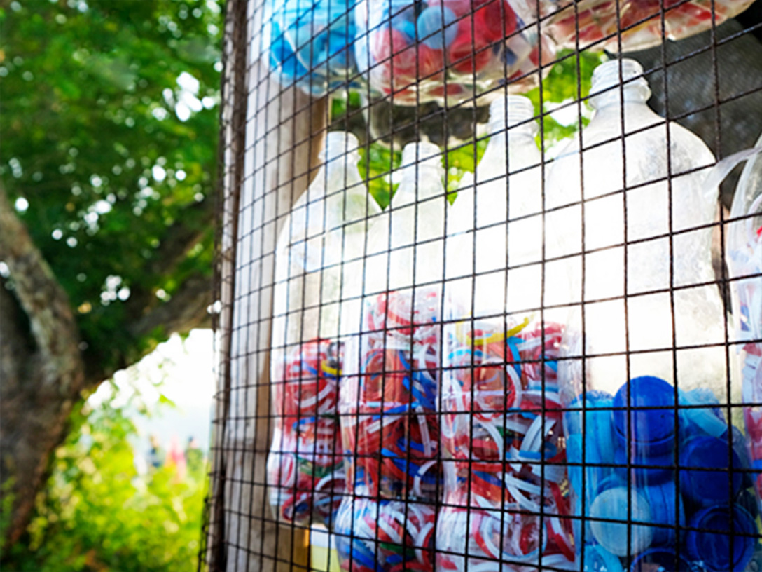 Several plastic bottles filled with caps, hanging from a grid with trees in the background
