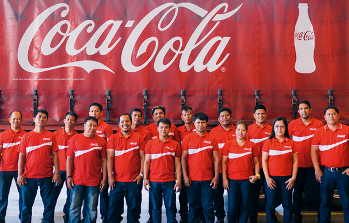 A group of people with Coca-Cola shirts smiling together in front of a Coca-Cola banner