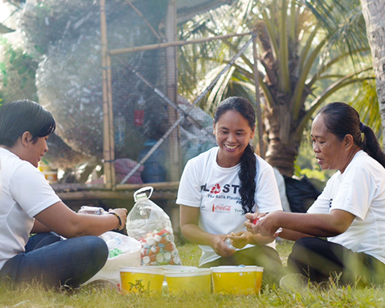 Three people sitted together on the grass collecting plastic bottle lids