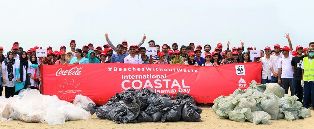 A large group of individuals on a beach holding a banner about 'International Coastal Cleanup Day', with many bags of collected waste in front of them