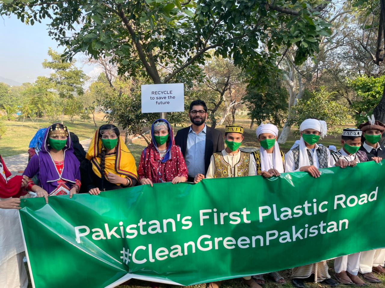 A group of people hold a banner with the phrase 'Pakistan's First Plastic Road #CleanGreenPakistan'