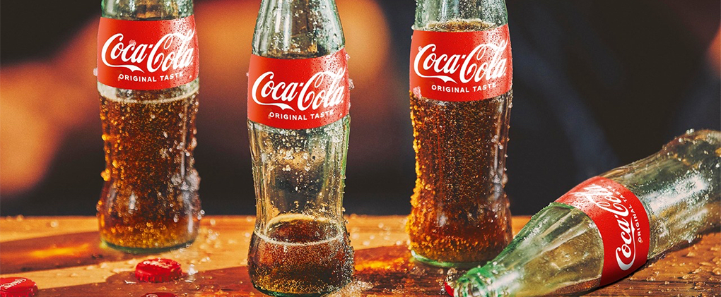 Front view of four open wet Coca-Cola Original Taste glass bottles on top of a wooden table.