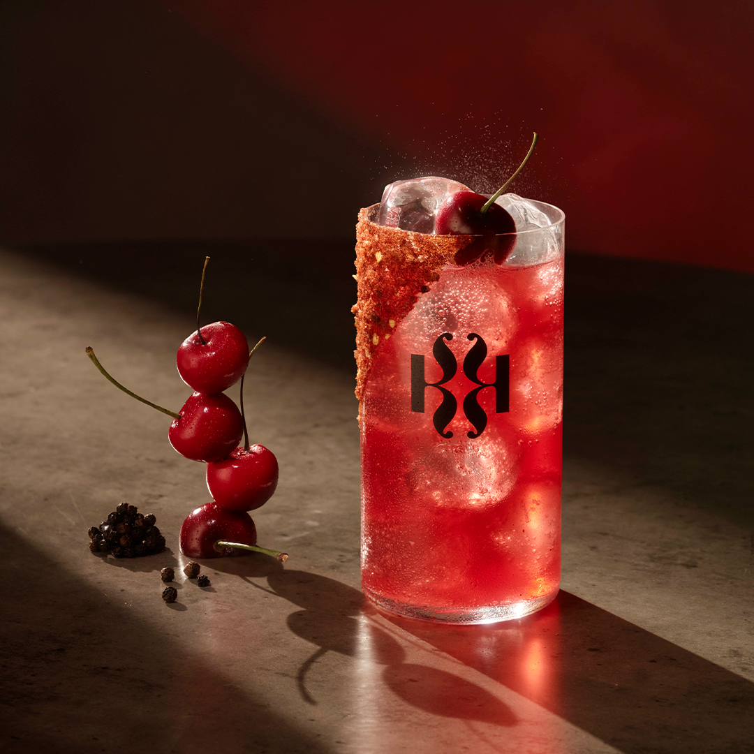 Czerwony drink z lodem i wiśnią w szklance Kinley na ciemnym tle.
